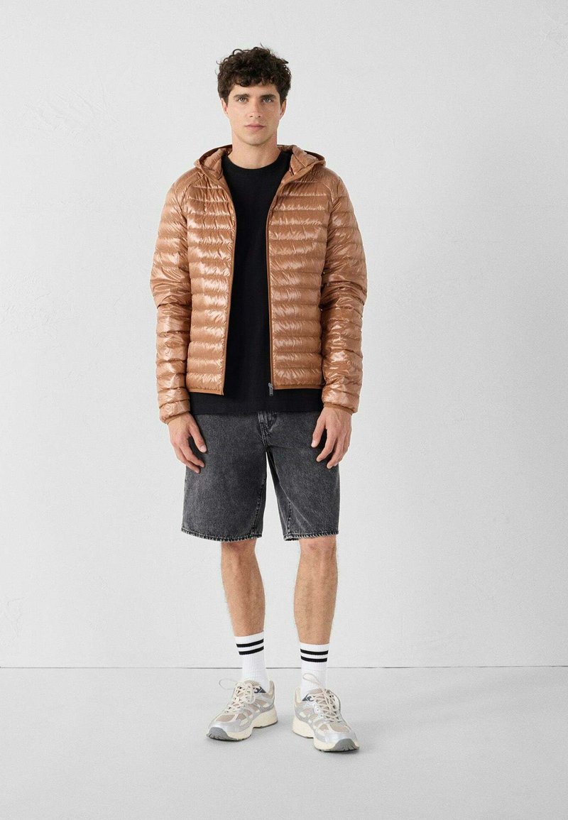 Giacca imbottita marrone con cappuccio, caratterizzata da una texture lucida e un motivo trapuntato, indossata sopra una maglia nera e abbinata a pantaloncini di denim e scarpe da ginnastica.