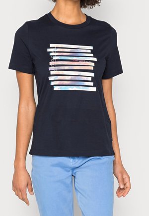 Person iført en sort t-shirt med hvide vandrette striber, der viser et mønster af himmel og skyer samt delvis tekst, kombineret med lyseblå bukser.