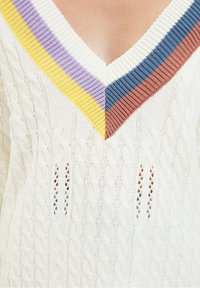 Pull en tricot blanc présentant un col en V profond avec des accents côtelés colorés en jaune, violet et marron. Texture en tricot torsadé avec de petits détails en œillets.