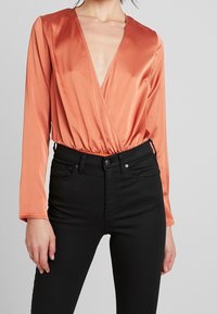 Langärmliger Body mit tiefem V-Ausschnitt aus satinorangem Stoff, mit einem gerafften Bund. Kombiniert mit hochgeschnittenen schwarzen Jeans.