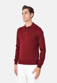 Polo a maniche lunghe bordeaux realizzato in tessuto morbido con colletto classico, patta con tre bottoni e polsini a costine. Pantaloni bianchi.