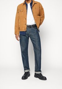 Chaqueta de pana marrón, camiseta blanca, camisa azul marino debajo, jeans de mezclilla azules, puños doblados, botas negras con suelas gruesas.