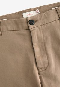 Beige cotton trousers featuring a button closure, zip fly, and subtle diagonal texture. Tag label visible near waistband.