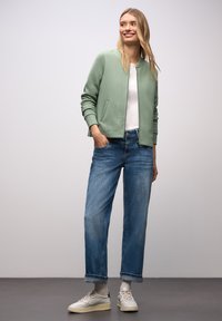 Bomber verde chiaro trapuntato, T-shirt bianca, jeans blu larghi e sneakers crema. Presenta materiale strutturato e vestibilità rilassata.