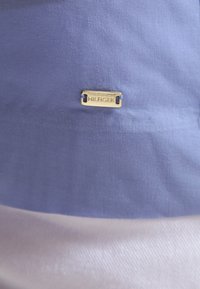 Sininen kangas, jossa on sileä pinta, keskellä kiinnitetty kultaiseen metallielementtiin kaiverrettu "HILFIGER"-teksti.