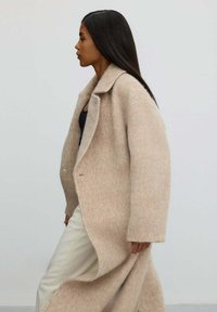 Žena s dlouhými tmavými vlasy, která má na sobě béžový volný vlněný kabát přes černý top a bílé kalhoty, kráčející před jednobarevným pozadím.