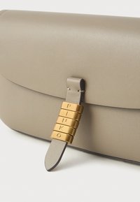 Bolso de mano de cuero beige con una textura suave, que cuenta con una correa de acento dorada con letras grabadas y un cierre de solapa.