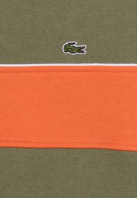 Tessuto verde con una striscia orizzontale arancione, piping bianco, e un piccolo logo di un coccodrillo verde sul lato sinistro.
