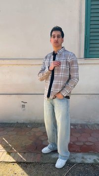 Jeune homme en chemise à carreaux claire, sous-vêtement noir, jean bleu clair et baskets blanches se tient à côté d'un mur pâle avec un volet de fenêtre vert.