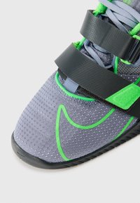Grå atletiske sko med grønne accenter, tekstureret stofoverdel og sorte velcro-stropper. Har en polstret tunge og fleksibel sål.