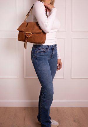 Frau trägt ein weißes langärmliges Oberteil und blaue Jeans, hält eine braune Wildleder-Handtasche über der Schulter und steht drinnen auf einem Holzboden.