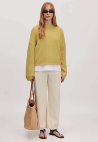 Gele, oversized gebreide trui, crèmekleurige broek, zwarte teenslippers en een geweven tote bag. Glad textuur met verlaagde schouders.