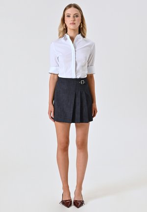Camicia bianca con maniche arrotolate abbinata a una gonna a pieghe scura con fibbia, indossata con tacchi a punta marroni.