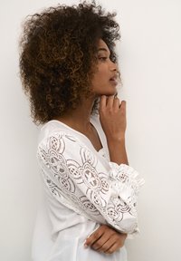 Weiße Bluse mit Spitzenverzierungen an den Ärmeln, die komplizierte Muster aufweist. Das Model hat lockiges Haar, der Stoff ist leicht strukturiert und der Schnitt ist lässig.
