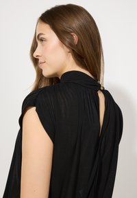 Schwarzes, leichtes Oberteil mit kurzen Ärmeln und hohem Kragen. Mit gerafftem Stoff und zwei dekorativen Knöpfen im Nacken. Glatte Textur.