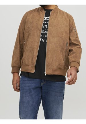 Uomo che indossa una giacca bomber in camoscio marrone sopra una t-shirt nera con stampa grafica e jeans blu su sfondo semplice.