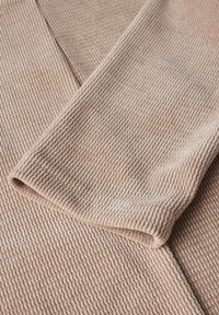 Tissu texturé de couleur beige clair avec un motif en relief, offrant une apparence douce et confortable. Les bords repliés montrent une finition lisse.
