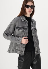 Gray denim jacket with classic collar, two front pockets, and button closure. Worn over a black textured top and paired with shiny black pants.