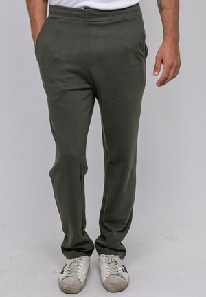 Homme portant un pantalon vert foncé et des baskets blanches, debout les mains dans les poches devant un fond clair uni. Seule la partie inférieure du corps est visible.