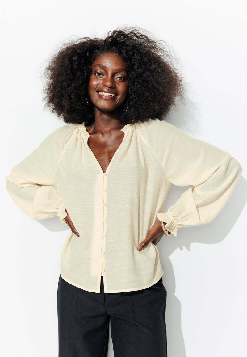 Blouse de couleur crème avec de longues manches bouffantes, décolleté en V, devant à boutons et détail de col froncé. Tissu texturé pour un aspect doux.