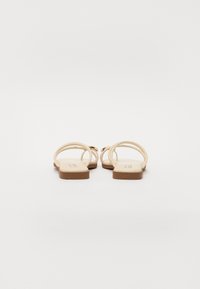 Beige flade sandaler med to buede stropper og en lille hæl. Sålen er glat, og stropperne har en blød tekstur, hvilket skaber et minimalistisk design.