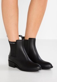 Black ankle rain boots with elastic side panels and low block heels, worn on bare legs against a white background.