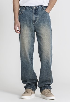 Homme portant un jean bleu délavé à coupe ample, des baskets beiges et une chemise blanche, une main dans la poche, sur un fond uni.