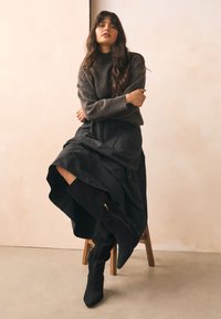 Dunkelgrauer Strickpullover, getragen mit einem schwarzen Maxirock mit hohem Schlitz, kombiniert mit schwarzen Wildleder-Stiefeln bis zum Knie. Hintergrund mit Holzhocker.