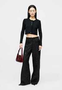 Schwarzer kurz geschnittener Pullover mit Knopfverschluss, kombiniert mit weit geschnittenen, schwarzen Hosen mit funkelnder Textur. Das Modell hält eine bordeauxfarbene Handtasche.