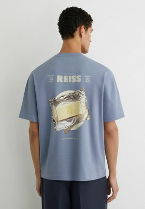 Muž oblečený v modrom tričku s potlačou masla na fólii a textom "REISS" na chrbte, stojaci pred jednoduchou svetlou stenou.