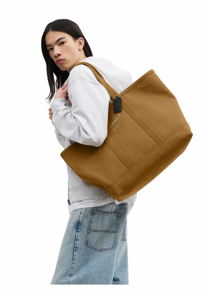 Personne portant un sweat à capuche blanc et un jean bleu clair tient un grand sac cabas marron Coach sur l'épaule sur fond blanc.
