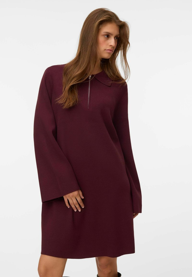 Femme portant une robe bordeaux foncé à manches longues avec un col et une fermeture éclair à l'avant, debout devant un fond blanc uni.