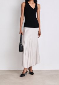 Donna che indossa un top senza maniche nero, una lunga gonna beige chiaro, tacchi a slingback neri e tiene una borsa nera a lato.