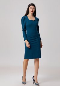 Robe ajustée teal avec un décolleté carré et des manches bouffantes. Fabriquée dans un tissu lisse, elle arrive aux hanches avec une coupe droite. Portée avec des chaussures à talons à motifs.