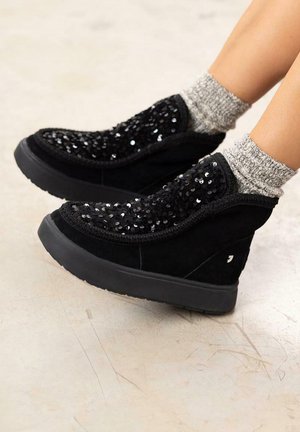 Des bottes en suède noir présentent un dessus en sequins avec une finition texturée, des panneaux latéraux élastiques et une semelle en caoutchouc plate, accompagnées de chaussettes en tricot gris.