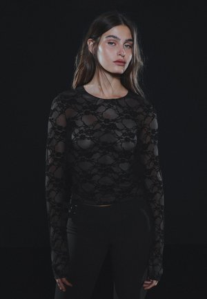 Une femme pose sur un fond sombre, vêtue d’un haut noir en dentelle transparente et d’un pantalon noir.