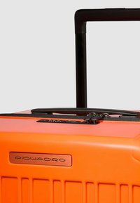 Orangener Hartschalenkoffer mit einem ausziehbaren schwarzen Griff, Reißverschluss und einem metallischen "Piquadro"-Logo. Strukturierte Oberfläche mit vertikalen Rillen.