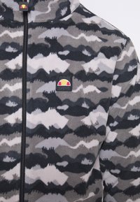Veste en polaire à motif camouflage gris et noir avec une fermeture éclair noire et un petit patch logo rectangulaire présentant un design rouge et jaune.
