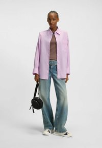 Camicia oversize button-up di un leggero viola sopra una maglietta marrone, abbinata a jeans a gamba larga scoloriti e sneakers bianche. Borsa a tracolla nera rotonda.