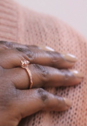 Anello in oro rosa con montatura a forma di corona, con una pietra centrale trasparente e piccoli accenti lungo la fascia. Texture liscia, design elegante.