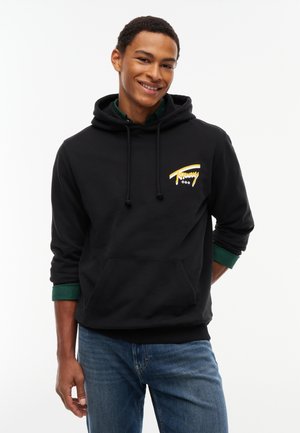 Ung mand smilende, iført en sort hættetrøje med et gult logo og blå jeans, står mod en ensfarvet hvid baggrund.