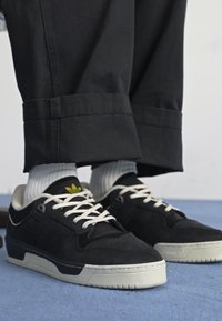 Melnas sporta kurpes ar zamšādu un ādas augšdaļu, baltām zoleēm un baltajiem šņorēm. Iezīmējas ar mazu dzeltenu logotipu uz mēles un teksturētām rakstu virsmām.