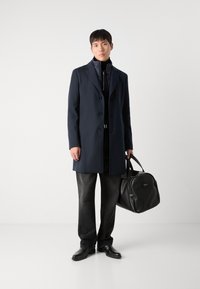 Capa de marinheiro com cinco botões, casaco interior preto com fecho éclair, jeans cinza escuro e mala de couro preta, finalizado com sapatos pretos.