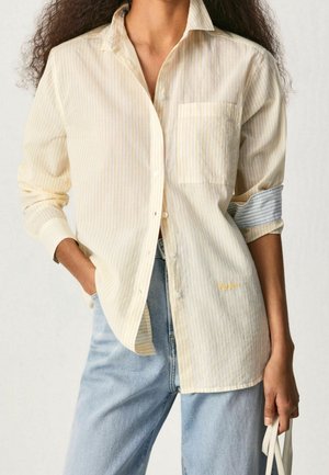 Persona con una camisa a rayas amarilla clara abotonada de forma holgada con las mangas remangadas y jeans azul claro, sosteniendo asas blancas de bolso con una mano.