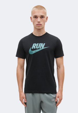 Jeune homme aux cheveux blonds courts portant un t-shirt noir Nike « RUN » et un short gris Nike, debout devant un fond clair uni.