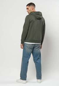 Mann von hinten, trägt eine olivgrüne Kapuzenjacke, weite blaue Jeans und weiße Turnschuhe vor einem schlichten hellen Hintergrund.