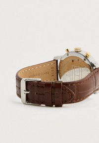 Καφέ δερμάτινο λουράκι ρολογιού με ασημένια αγκράφα και δύο θηλιές, συνδεδεμένο με μια ασημένια κάσα ρολογιού με χρυσό κορώνα και κουμπιά.