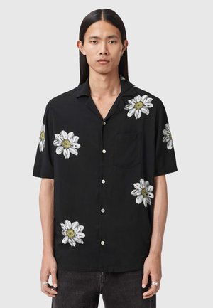 Ung person med langt hår iført en sort kortærmet skjorte med hvide marguritblomster og mørke bukser, stående mod en ensfarvet baggrund.