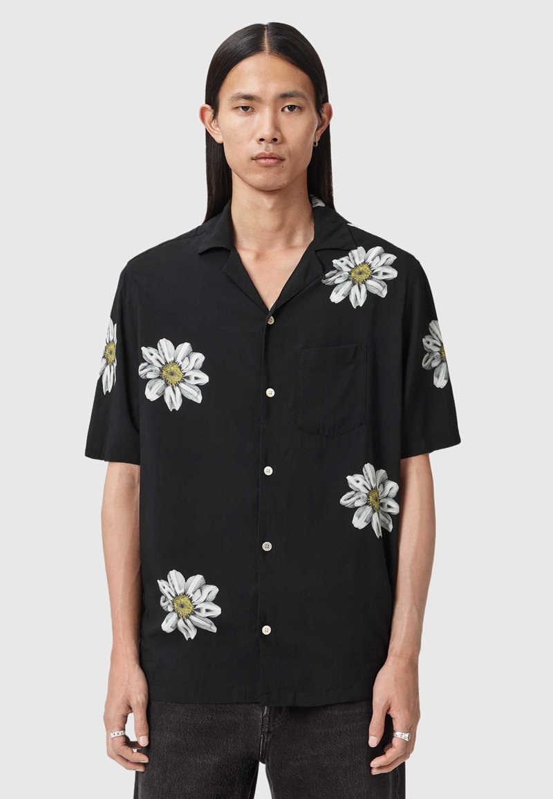 Młoda osoba z długimi włosami nosi czarną, krótkorękawową koszulę na guziki z białymi nadrukami stokrotek oraz ciemne spodnie, stoi na tle jednolitego tła.