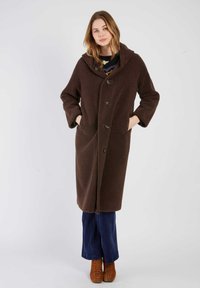 Długi brązowy płaszcz z polaru z kapturem, z dużymi guzikami, bocznymi kieszeniami i luźnym krojem. Nosi się go z granatowymi spodniami sztruksowymi i brązowymi butami.